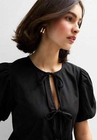 Blusa negra con mangas cortas abullonadas, escote atado y una textura suave. Presenta un corte holgado y sutiles detalles elegantes.