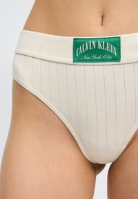 Μπεζ ριμπε ανώτερου κοψίματος εσώρουχο με πράσινο λογότυπο Calvin Klein New York City, φορεμένο από ένα άτομο, που απεικονίζεται από τη μέση έως τον άνω μηρό.