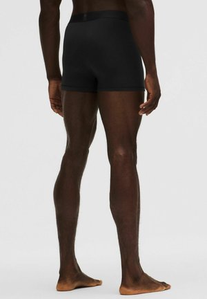 Tagantvaate seisvast isikust, kes kannab musta keha järgivaid bokserpükse ning on paljajalu, käed lõdvalt piki keha.