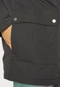 Giacca leggera nera presenta una grande tasca frontale con chiusura a scatto, texture visibile e cuciture a contrasto.