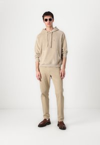 Hanorac din bumbac bej cu buzunar în față, asociat cu pantaloni bej deschis. Modelul poartă ochelari de soare negri și pantofi din piele maro.