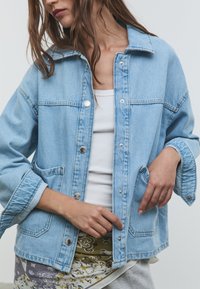 Svetlo modra denim jakna s sproščenim krojem, opremljena s pritisnimi gumbi, dvema sprednjima žepoma in klasičnim ovratnikom. Mehka tekstura in strukturiran dizajn.