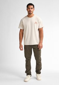 Beige katoenen t-shirt met een kleine opdruk op de linkerborst, gecombineerd met olijfgroene cargobroeken met meerdere zakken en witte sneakers.