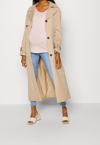 Trench beige con cierre de botones, corte holgado y bolsillos laterales, llevado sobre una blusa a rayas rosa y jeans azules, combinado con mules de tacón cuadrado blancos.