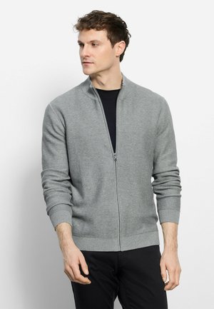 Mann trägt einen grauen, gerippten Reißverschluss-Pullover über einem schwarzen Hemd, schaut nach links, die Hände entspannt an den Seiten.