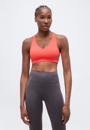 Femme en forme aux cheveux tressés portant un soutien-gorge de sport corail et un legging gris foncé taille haute, debout devant un fond clair uni.