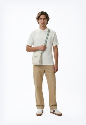 Muž v bílém texturovaném polo tričku s krátkým rukávem, béžových kalhotách, bílých teniskách a malé béžové kabelce přes rameno se zeleným popruhem a logem Calvin Klein.
