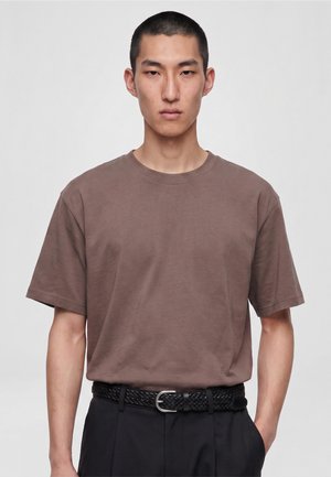 Jeune homme aux cheveux courts portant un t-shirt marron rentré dans un pantalon noir avec une ceinture noire tressée, debout devant un fond uni.