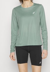 Hellgrünes langärmliges Sportshirt mit Rundhalsausschnitt und Logo. Kombiniert mit enganliegenden schwarzen Shorts. Glatte Textur, minimales Design.