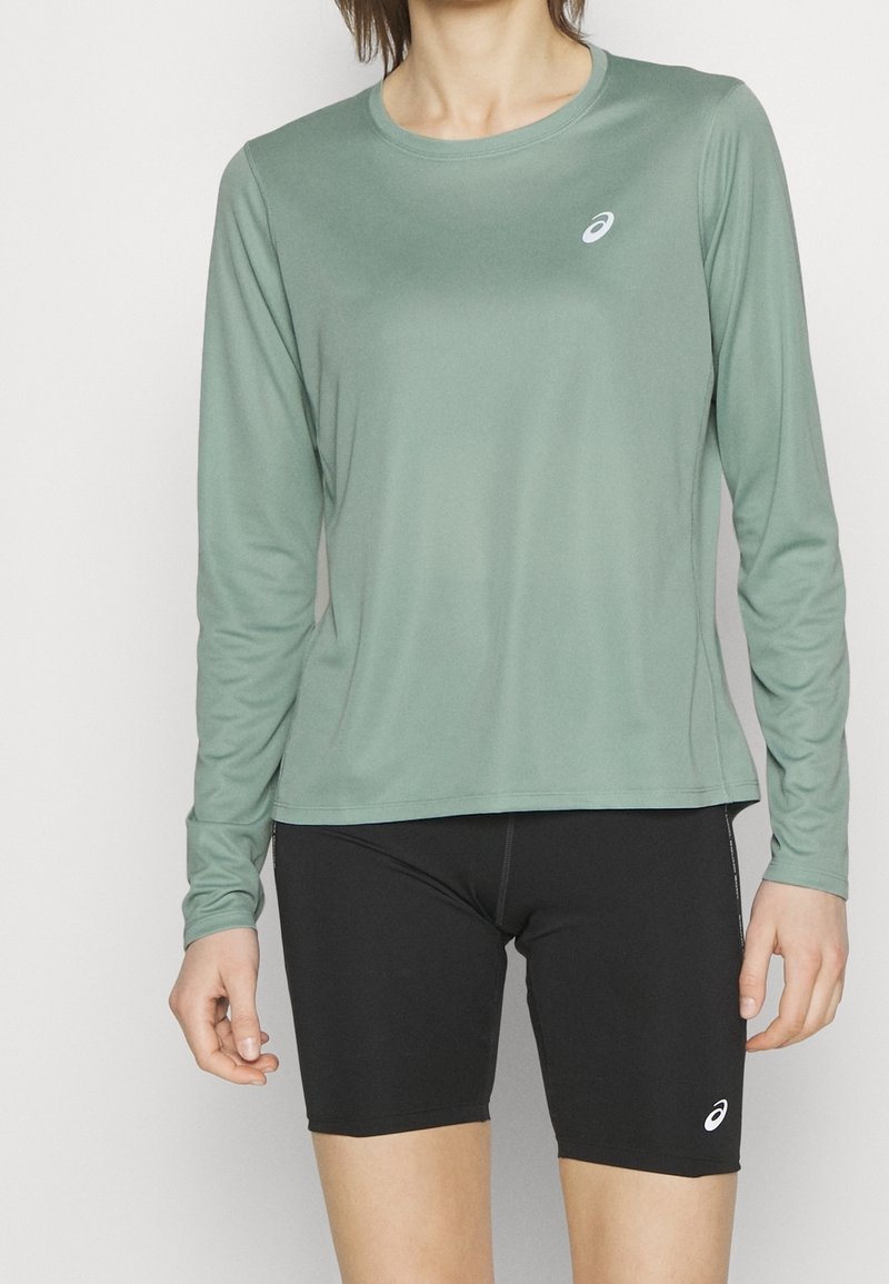 Hellgrünes langärmliges Sportshirt mit Rundhalsausschnitt und Logo. Kombiniert mit enganliegenden schwarzen Shorts. Glatte Textur, minimales Design.