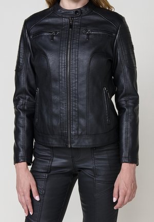 Woman wearing black leather jacket and matching black leather pants with zipper and stitching details, standing against gray background.