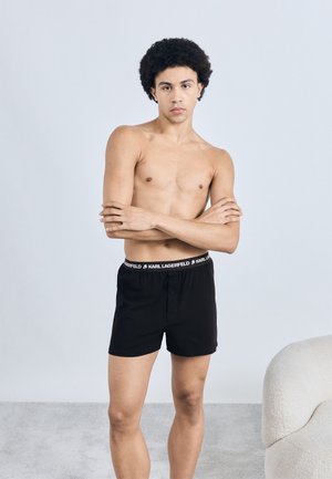 Black boxer shorts featuring a branded waistband. The material appears smooth, with a button closure at the front and a relaxed fit design.