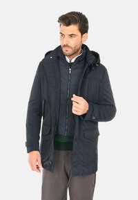 Giacca esterna blu navy con cappuccio, dotata di chiusura frontale con cerniera e due grandi tasche. Lo strato interno è un gilet trapuntato. Tessuto morbido e liscio.