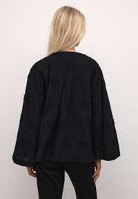 Blusa nera ricamata con maniche lunghe, caratterizzata da motivi floreali e una vestibilità ampia, realizzata in tessuto testurizzato.