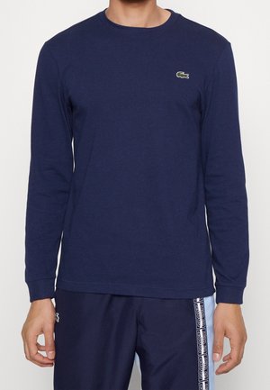 Mann trägt ein marineblaues Langarmshirt mit kleinem grünem Krokodil-Logo und passende marineblaue Hose mit weißem Streifen-Detail.