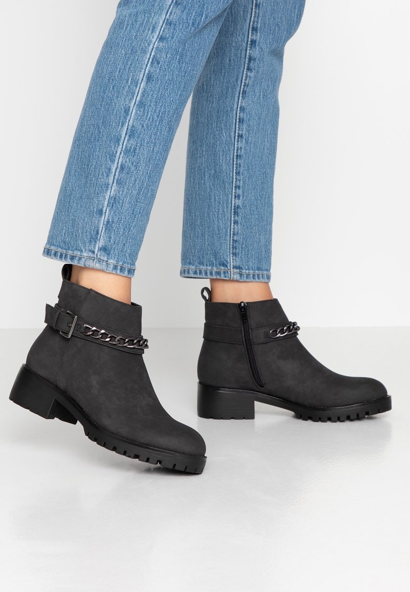 Personne portant des bottines noires avec des détails en chaîne et des fermetures éclair latérales, associées à un jean bleu raccourci sur un sol clair.