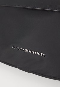 Sac en nylon noir avec une texture lisse, fermeture éclair et logo argenté de Tommy Hilfiger en police moderne. Design minimaliste.
