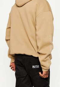 Felpa con cappuccio beige a maniche lunghe, con logo bianco sulla manica sinistra, abbinata a pantaloni cargo neri con una toppa del logo.