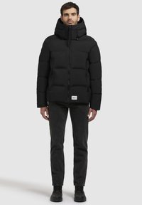 Chaqueta de plumas negra con cuello alto, cierre de cremallera, bolsillos laterales y costuras visibles. Usada sobre unos vaqueros oscuros y botas negras.