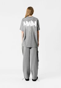 Camiseta gris de gran tamaño con un diseño de goteo blanco en la parte posterior, combinada con pantalones de chándal gris de pierna ancha. El material parece suave y casual.
