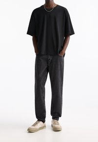 Maglietta nera oversize, vestibilità comoda, abbinata a jeans denim grigio scuro. Il modello indossa sneakers beige e grigie. Semplice catena d'argento al collo.