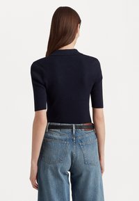 Kvinna med långt brunt hår som bär en svart kortärmad ribbad topp och högmidjade blå jeans med ett svart bälte, vänd bort.