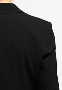 Černý blazer s hladkou texturou a strukturovanými ramenními vycpávkami. Obsahuje klopový límec a čisté švy podél švů.