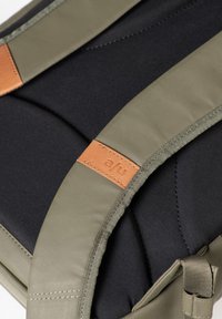 Lo zaino verde oliva presenta bordi imbottiti, tessuto nero e dettagli in pelle marrone con cuciture visibili e logo.