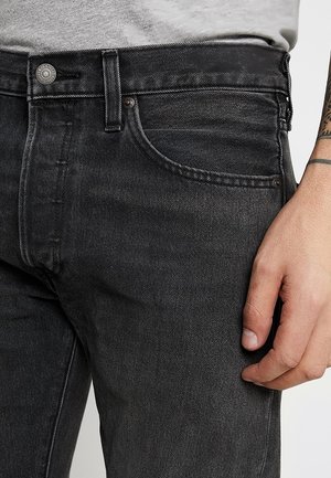 Close-up of a person wearing black denim jeans with a visible front pocket, button, and tattooed forearm resting beside the hip.