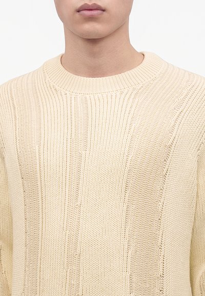 Buste masculin portant un pull en maille côtelée crème à col rond sur un fond uni.