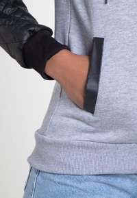 Mano en el bolsillo de una sudadera gris con ribete negro, lleva puesta una manga de chaqueta acolchada negra y jeans azul claro.