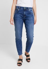 Tmavě modré džíny z denimu s úzkým střihem, pěti kapsami a knoflíkovým zapínáním. Nosené s černými koženými sandály s širokým páskem a cvočky.