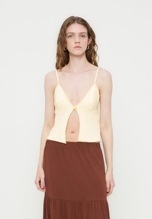 Jeune femme aux cheveux bruns ondulés portant un haut crème sans manches avec une ouverture devant et une jupe marron taille haute sur un fond uni.