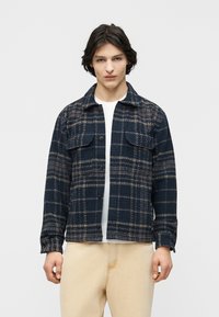 Νεαρός άνδρας που φοράει μπλε ναυτικό και μπεζ καρό πουκάμισο με κουμπιά πάνω από λευκή μπλούζα και μπεζ παντελόνια, στέκεται μπροστά σε ένα απλό φωτεινό φόντο.