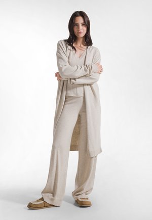 Kvinde stående med armene krydset iført beige strikket cardigan, matchende vidde bukser og brune tøfler på en ensfarvet hvid baggrund.