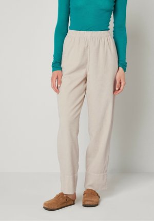 Kobieta ubrana w beżowe spodnie sztruksowe, turkusową bluzkę z długim rękawem i brązowe buty wsuwane, stojąca na tle jednolitego tła.