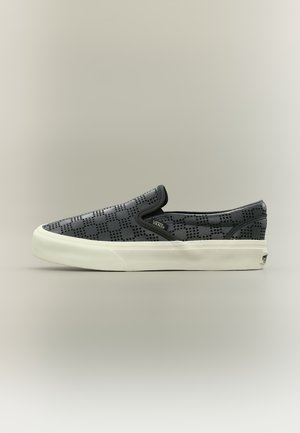 Chaussure Vans noire slip-on avec motif à grille perforée et semelle en caoutchouc blanche sur un fond clair neutre.