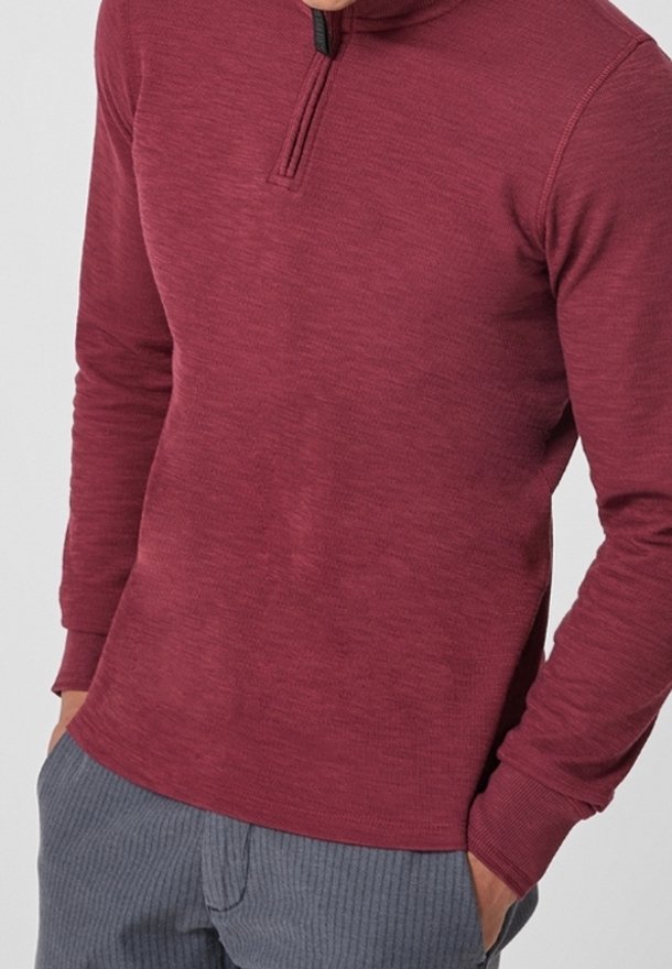 Mann trägt einen roten Langarmpullover mit einem Viertel-Reißverschluss und grauen Ripp-Hosen, die Hände in den Taschen, vor einem schlichten Hintergrund.
