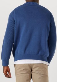 Maglione blu lavorato a maglia con texture a coste, scollo rotondo e vestibilità rilassata. Indossato sopra una camicia bianca e abbinato a pantaloni chiari.