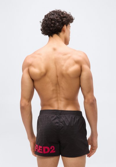 Homme musclé aux cheveux bouclés et foncés, debout torse nu de dos, portant un short de bain noir avec le texte rouge "RED2" sur la jambe gauche.