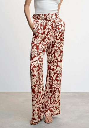 Vrouw draagt een hooggetailleerde, wijde broek met een rood-beige bloemmotief, gecombineerd met een wit mouwloos geribd shirt en sandalen.