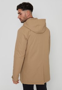 Beige gewatteerde jas met een gladde textuur, ruime pasvorm en zichtbare stikseldetails. Heeft een rechte rugontwerp en verstelbare manchetten.