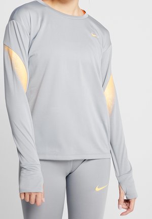 Osoba ubrana w jasno szary, sportowy top z długim rękawem i legginsy z małymi żółtymi logo Nike Swoosh na klatce piersiowej, rękawie i udzie.
