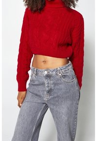 Roter kurz geschnittener Pullover mit Zopfmuster und Rollkragen. Paar hellgraue Jeans mit hoher Taille, leicht lockerer Passform, mit klassischen Taschen.