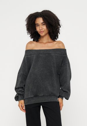 Femme aux cheveux bouclés portant un pull oversize gris foncé décolleté aux épaules et un pantalon noir, debout devant un fond clair uni.
