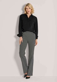 Vrouw met kort blond haar, gekleed in een zwarte button-up shirt, zwarte en witte geblokte broek en zwarte puntige schoenen, staand tegen een muur.