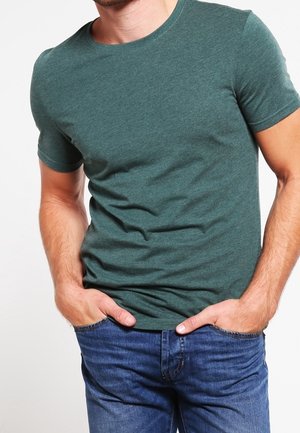 Mężczyzna w dopasowanej zielonej koszulce z krótkim rękawem i niebieskich dżinsach, stojący z rękami w kieszeniach na jednolitym jasnym tle.