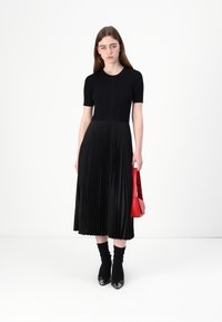 Vestito nero a pieghe con maniche corte, top in maglia testurizzata, gonna fluida, abbinato a una borsa rossa e tacchi neri a punta.