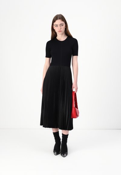 Robe noire plissée avec des manches courtes, haut en tricot texturé, jupe fluide, assortie à un sac à main rouge et des escarpins noirs à talons pointus.