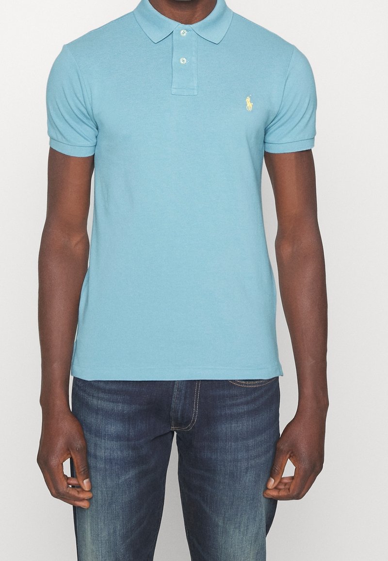 Homme portant un polo à manches courtes bleu clair avec un petit logo jaune sur la poitrine et un jean bleu foncé délavé, sur fond blanc.